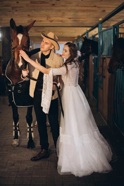 Handsome young cowboy man and his charming boho bride pose in a stable with horses