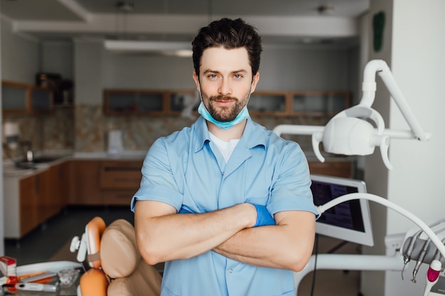 Handsome young dentist in white coat is looking at camera and smiling while standing with crossed arms in his office.