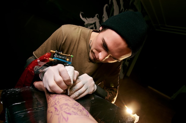 Handsome young guy in a black hat and with tattoos, beats a tattoo on his arm, tattoo salon