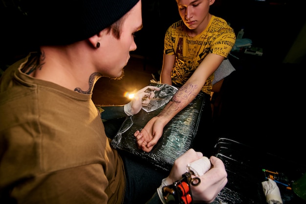 Handsome young guy in a black hat and with tattoos, beats a tattoo on his arm, tattoo salon