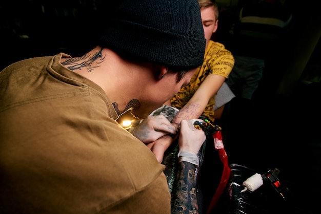 Handsome young guy in a black hat and with tattoos, beats a tattoo on his arm, tattoo salon