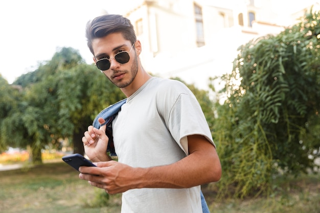 Handsome young guy walking outdoors listening to music at the street using mobile phone chatting looking camera