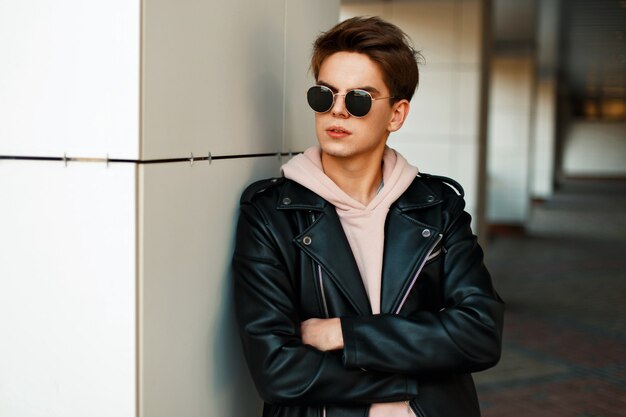 Handsome young guy with sunglasses in a fashionable branded jacket stands near a white wall