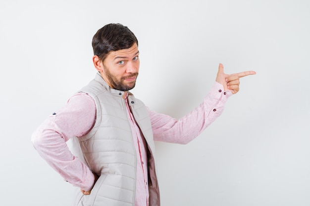 Handsome young male in shirt, vest pointing right, with hand on waist and looking confident , front view.