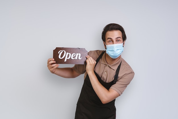 Handsome young man in apron wearing protective face mask and holding open sign while standing agains...