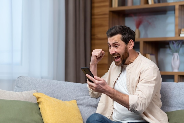 A handsome young man in a beige shirt is sitting on the sofa at home and playing games on the phone