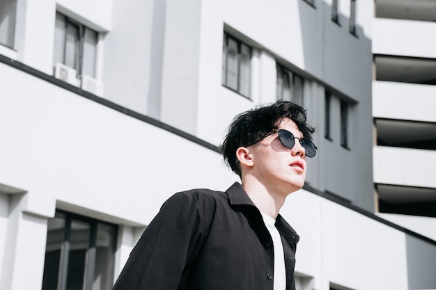 Handsome young man in a black shirt standing against the backdrop of city buildings and looking away through black sunglasses