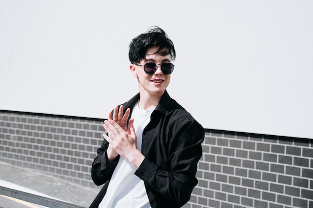 Handsome young man in a black shirt standing against the backdrop of city buildings and looking away through black sunglasses