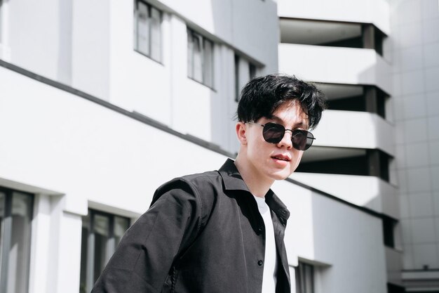 Handsome young man in a black shirt standing against the backdrop of city buildings and looking through black sunglasses