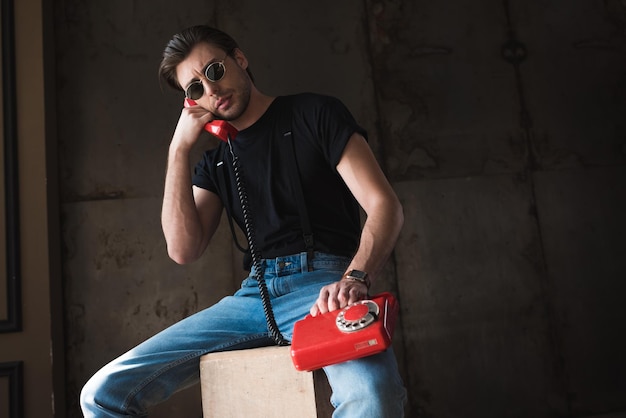 Handsome young man in black tshirt and sunglasses talking by retro wired red phone