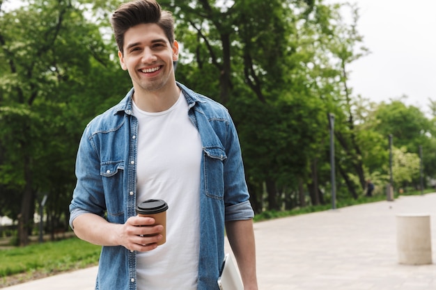 handsome young man in casual clothes drinking takeaway coffee and holding laptop while walking through green park