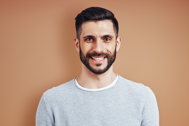 Handsome young man in casual wear smiling and looking at camera while standing against brown wall