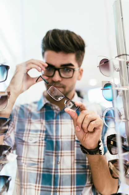 Handsome young man choosing eyeglasses frame in optical store.