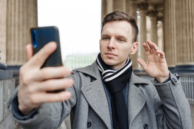 Handsome young man in coat walking on street holds mobile phone and shoots