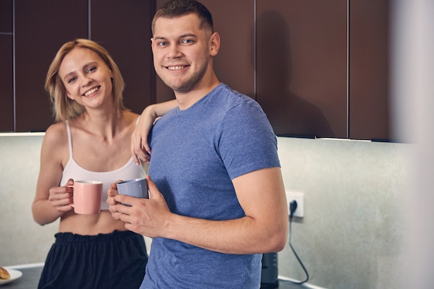 Handsome young man and cute blond woman smiling while relaxing in kitchen with cup of tea