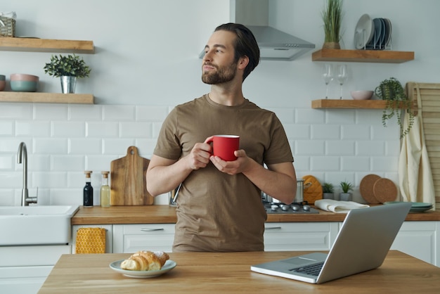 Handsome young man enjoying coffee and looking thoughtful while spending time at the kitchen