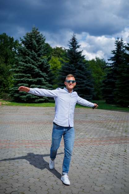 A handsome young man of European appearance wearing sunglasses is dressed in a shirt and jeans. The guy walks down the street, he is stylishly dressed