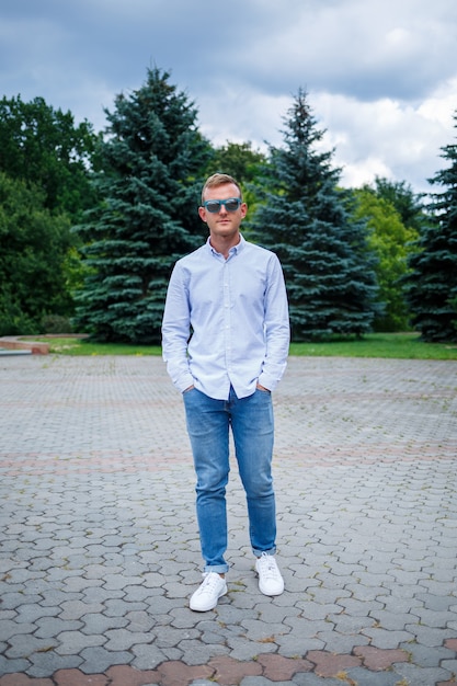 A handsome young man of European appearance wearing sunglasses is dressed in a shirt and jeans. The guy walks down the street, he is stylishly dressed