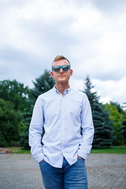 A handsome young man of European appearance wearing sunglasses is dressed in a shirt and jeans. The guy walks down the street, he is stylishly dressed