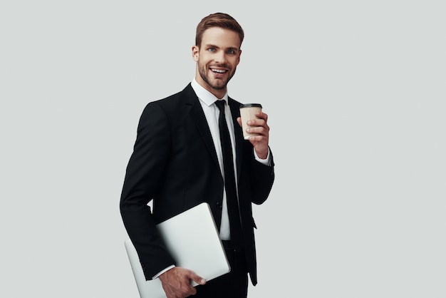 Handsome young man in formalwear carrying laptop and looking at camera with smile while standing against grey background