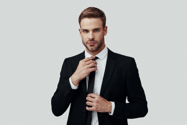 Handsome young man in formalwear looking at camera and adjusting tie while standing against grey background