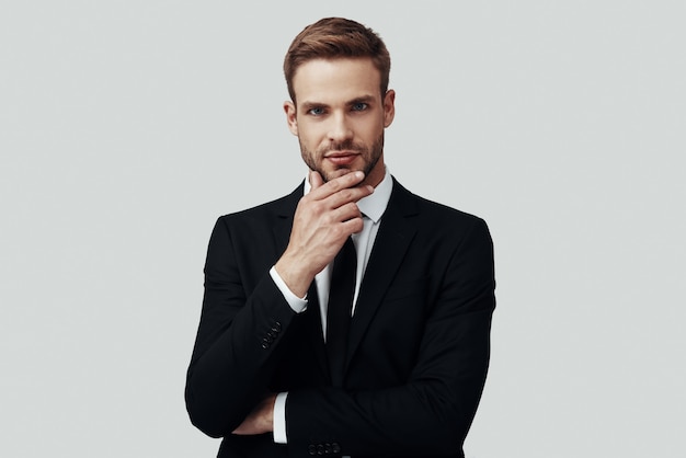 Handsome young man in formalwear looking at camera and keeping hand on chin while standing against grey background