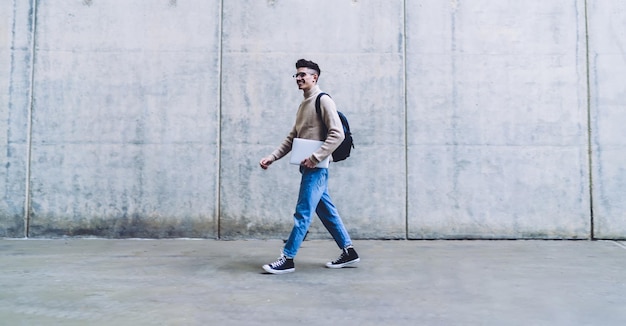 handsome young man in glasses and sweater with backpack holding white laptop while smiling