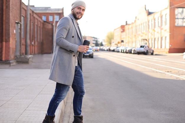 Handsome young man in grey coat and hat crossing the street with a cup of coffee.