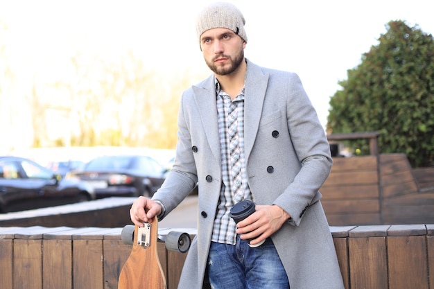 Handsome young man in grey coat and hat drinking coffee, resting, standing with longboard. Urban skateboarding concept.