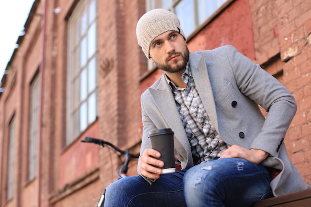 Handsome young man in grey coat and hat sitting on a bench relaxed drinking coffee and thinking near his bicycle.