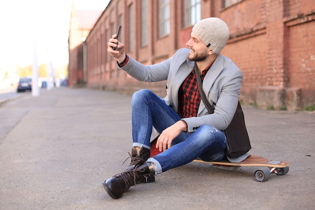 Handsome young man in grey coat and hat sitting on the longboard and taking selfie on the street in the city.