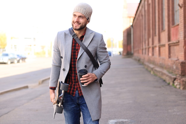 Handsome young man in grey coat and hat walking on the street, using longboard.