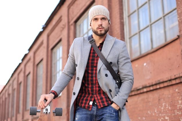 Handsome young man in grey coat and hat walking on the street, using longboard.