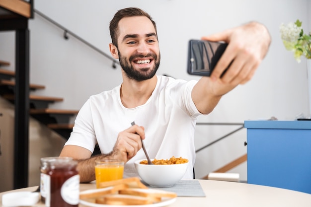 Handsome young man having breakfast while sitting at the kitchen, taking a selfie