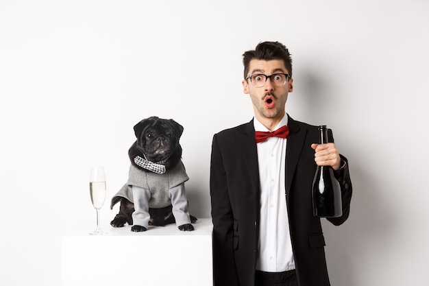 Handsome young man and his puppy celebrating New Year holiday, black pug and dog owner standing in suits, guy holding champagne, white.