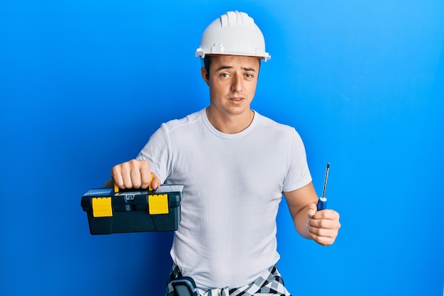 Handsome young man holding toolbox and screwdriver clueless and confused expression doubt concept