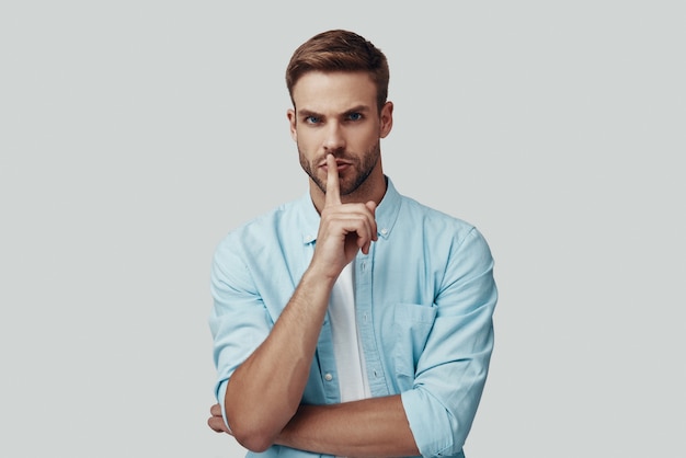 Handsome young man looking at camera and keeping finger on lips while standing against grey background