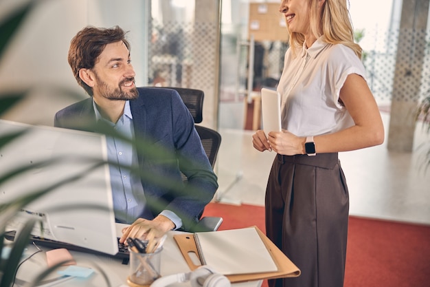 Handsome young man looking at charming lady and smiling while sitting at the table with computer in office