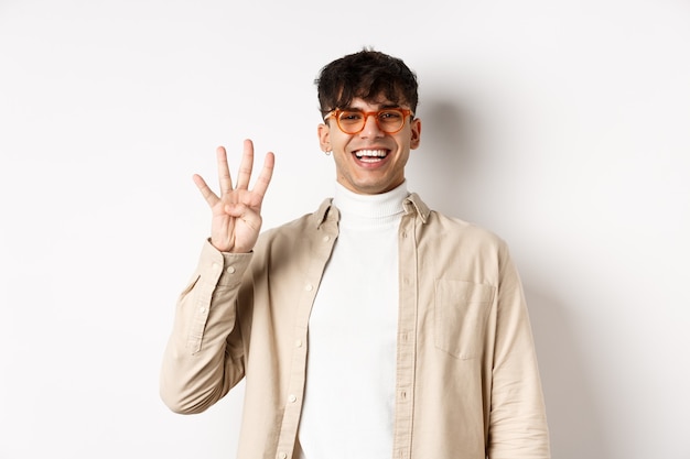 Handsome young man making order, showing number four fingers and smiling, standing on white wall.