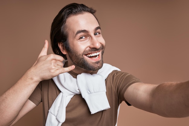 Handsome young man making selfie and gesturing while standing against brown background