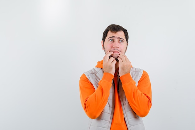 Handsome young man in orange hoodie and white vest put fingers about mouth and full of fear isolated on white background