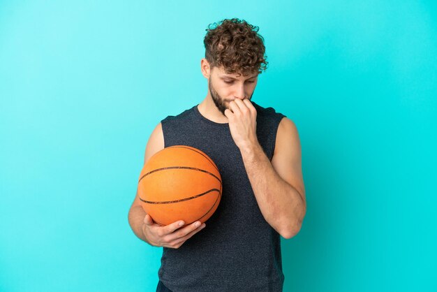 Handsome young man playing basketball isolated on blue background having doubts