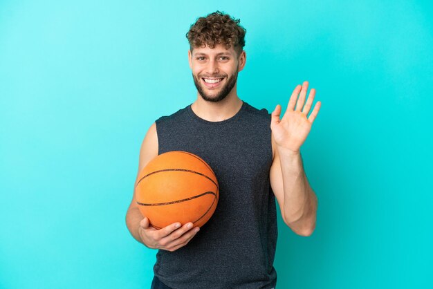 Handsome young man playing basketball isolated on blue background saluting with hand with happy expression