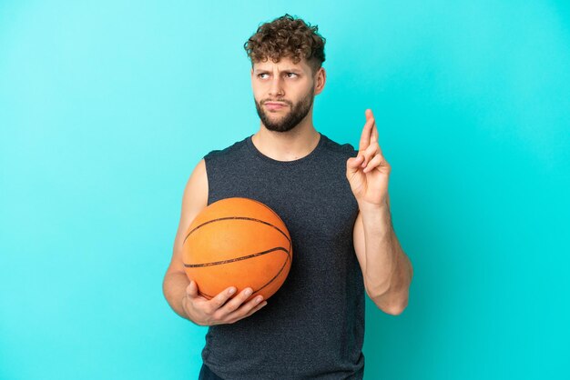 Handsome young man playing basketball isolated on blue background with fingers crossing and wishing the best