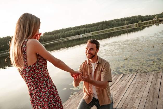 Handsome young man putting engagement ring on hand of beloved pretty girlfriend, asking to marry. Guy standing on his knee during sunset offering engagement ring to excited woman on lake background