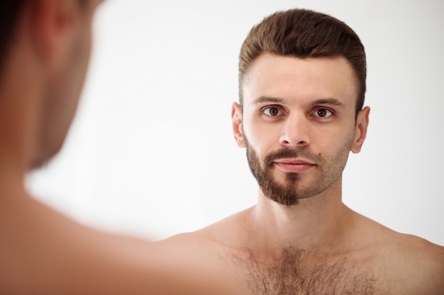 Handsome young man shaving his beard in the bathroom. Portrait of a stylish naked bearded man examining his face in-home mirror.