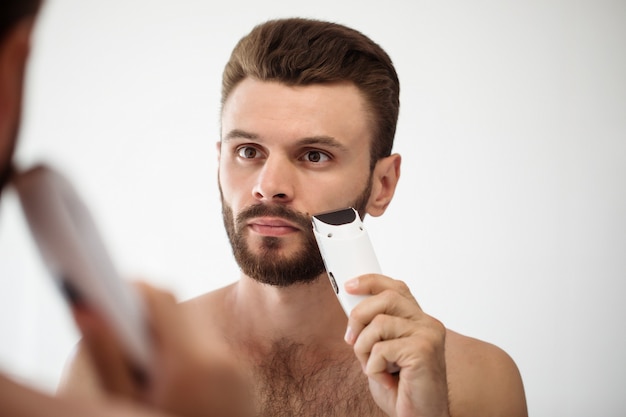 Handsome young man shaving his beard in the bathroom. Portrait of a stylish naked bearded man examining his face in-home mirror.