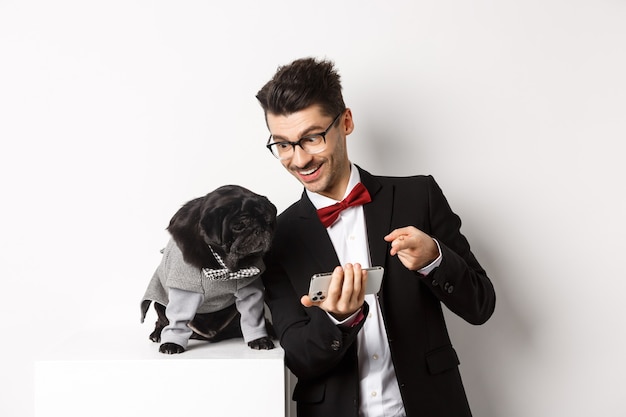 Handsome young man showing something on mobile phone to his dog. Owner shopping online with pet, standing in costumes over white.