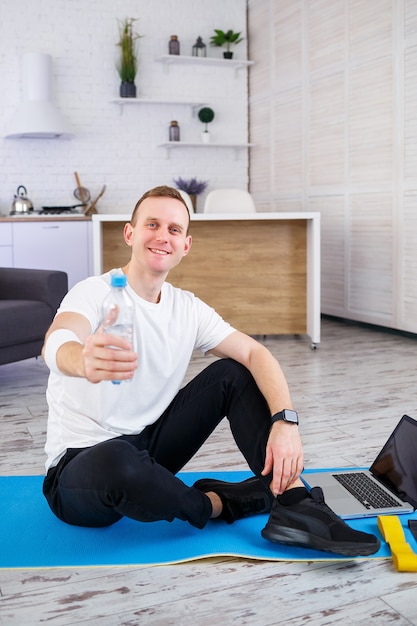 Handsome young man sitting on the floor and holding bottle of water while having online workout