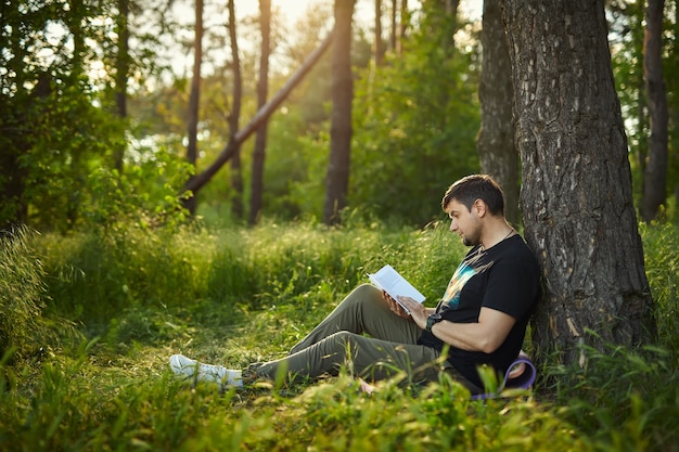 Handsome young man sitting in the forest on the grass, leaning against a tree, reading a book